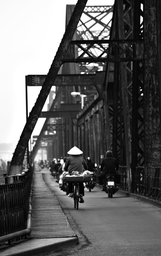 Black and white of native people riding bicycles and scooters on asphalt bridge with metal constructions and fence in town