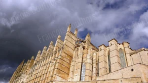 Photographie d’architecture d’une cathédrale gothique éclairée par la lumière, contrastant avec un ciel dramatique chargé de nuages.