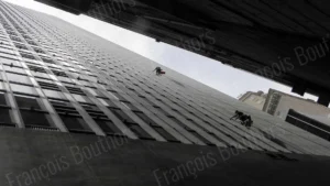Travailleurs suspendus nettoyant la façade d’un gratte-ciel, vue en contre-plongée entre deux immeubles. Une photographie d'architecture en noir et blanc.