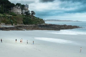 Maison en bord de falaise en Bretagne surplombant une grande plage blanche avec des personnes au loin, sous un ciel orageux – photographie François Bouthors