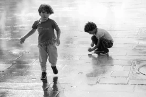Photo de rue en noir et blanc d’enfants jouant sur une place humide avec jets d’eau – François Bouthors