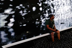 Street photo d’un homme assis face à une projection lumineuse abstraite dans un espace urbain – François Bouthors