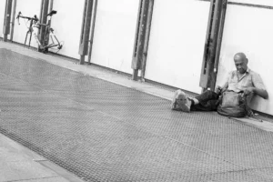 Street photo noir et blanc d’un homme allongé contre un mur urbain avec son sac et un vélo à proximité – François Bouthors